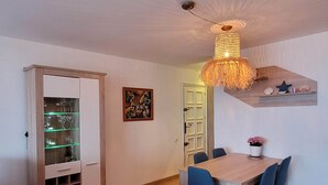 En matplats med ett träbord och blå stolar, ett glasskåp med vinglas, en hängande lampa och en vägghylla med dekorativa föremål.