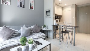 Ein modernes Wohnzimmer mit grauem Sofa, einem Couchtisch mit Dekoration, einem kleinen Esstisch mit Stühlen und einem durch eine offene Tür einsehbarer Küchenbereich.