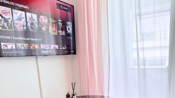 A modern dining area with a round table, two plush pink chairs, a remote control, and a decorative vase. A large screen displaying movie highlights is mounted on the wall. - Charming apartment | balcony, close to the center, self check-in (Augsburg)