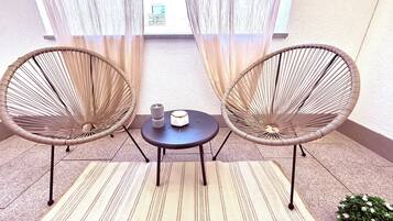 Two wicker chairs, a small round table, and a candle on a rug in a sunlit room with a view through sheer curtains.