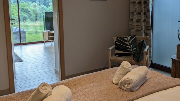 A bedroom with a bed, two rolled towels, a framed sign on the wall, a chair, and a view of the outdoors through a door. - Aux Fleurs des Champs (Lautenbach)