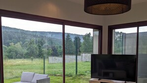 A modern living room with a large TV, comfortable seating, and a view of a grassy area through large windows. - Aux Fleurs des Champs (Lautenbach)