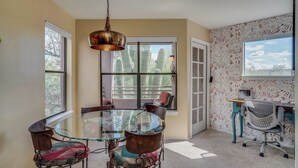 Un espace repas avec une table en verre et des chaises en métal, un luminaire suspendu et une vue sur des cactus à travers de grandes fenêtres.