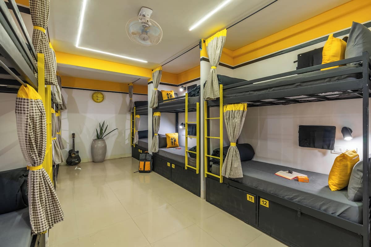 A dormitory room with bunk beds, checkered curtains, a fan, a guitar, and a potted plant.