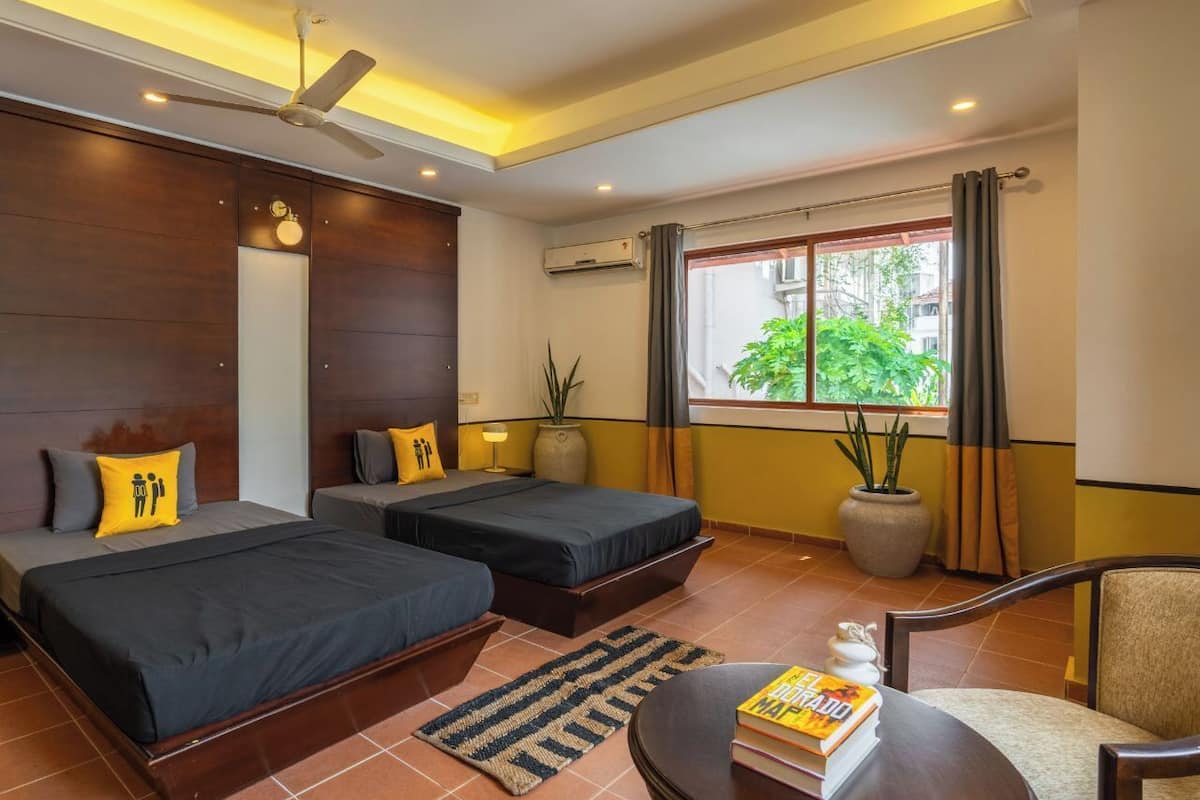 A hotel room with two beds, a ceiling fan, an air conditioning unit, a window with curtains, and a table with books.
