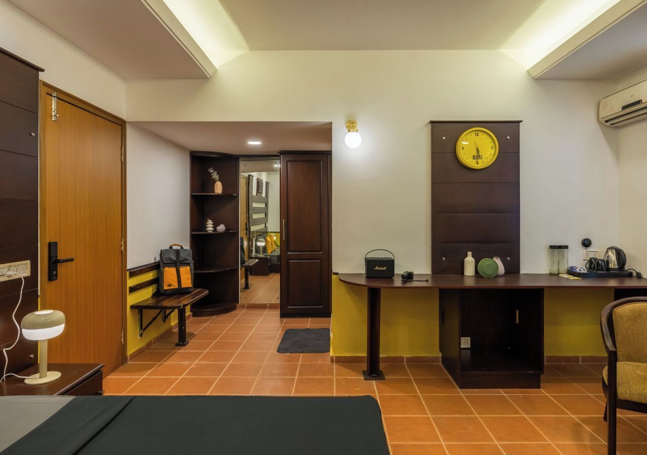 Un espacio habitable pequeño con una puerta de madera, un banco, un escritorio con un reloj y una pequeña zona de kitchenette.