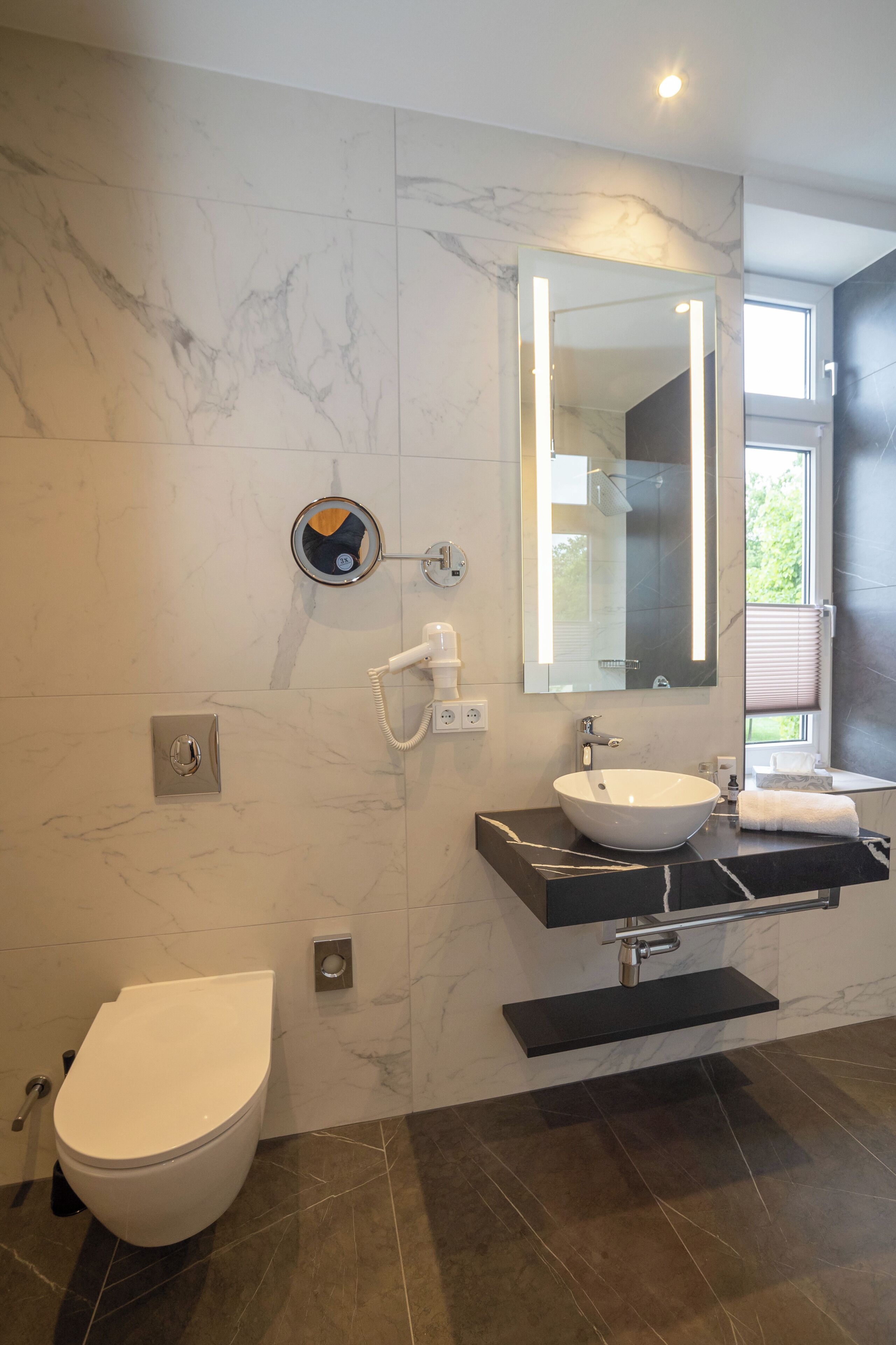 A modern bathroom with a white toilet, a sink with a mirror above, and a window with blinds.