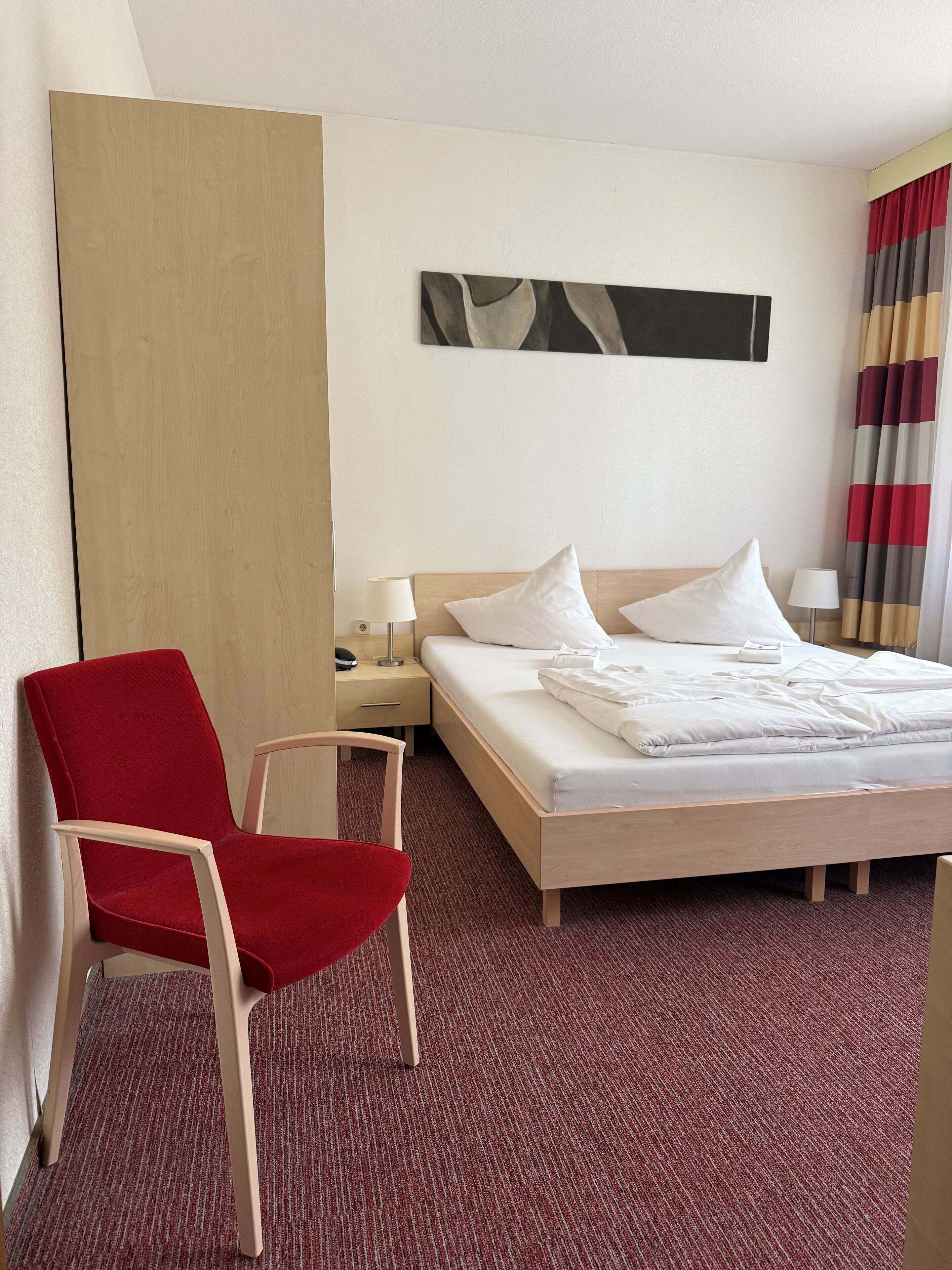 Ein Hotelzimmer mit Bett, einem roten Sessel, einem Nachttisch, einer Lampe und einem Wandbild.