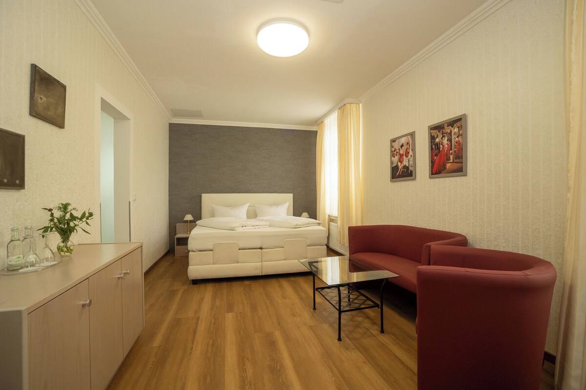 Habitación de hotel con cama, un sillón rojo, una mesita, una mesita de noche y televisor montado en la pared.