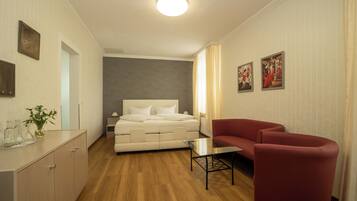 A hotel room with a bed, a red armchair, a small table, a nightstand, and a wall-mounted TV.