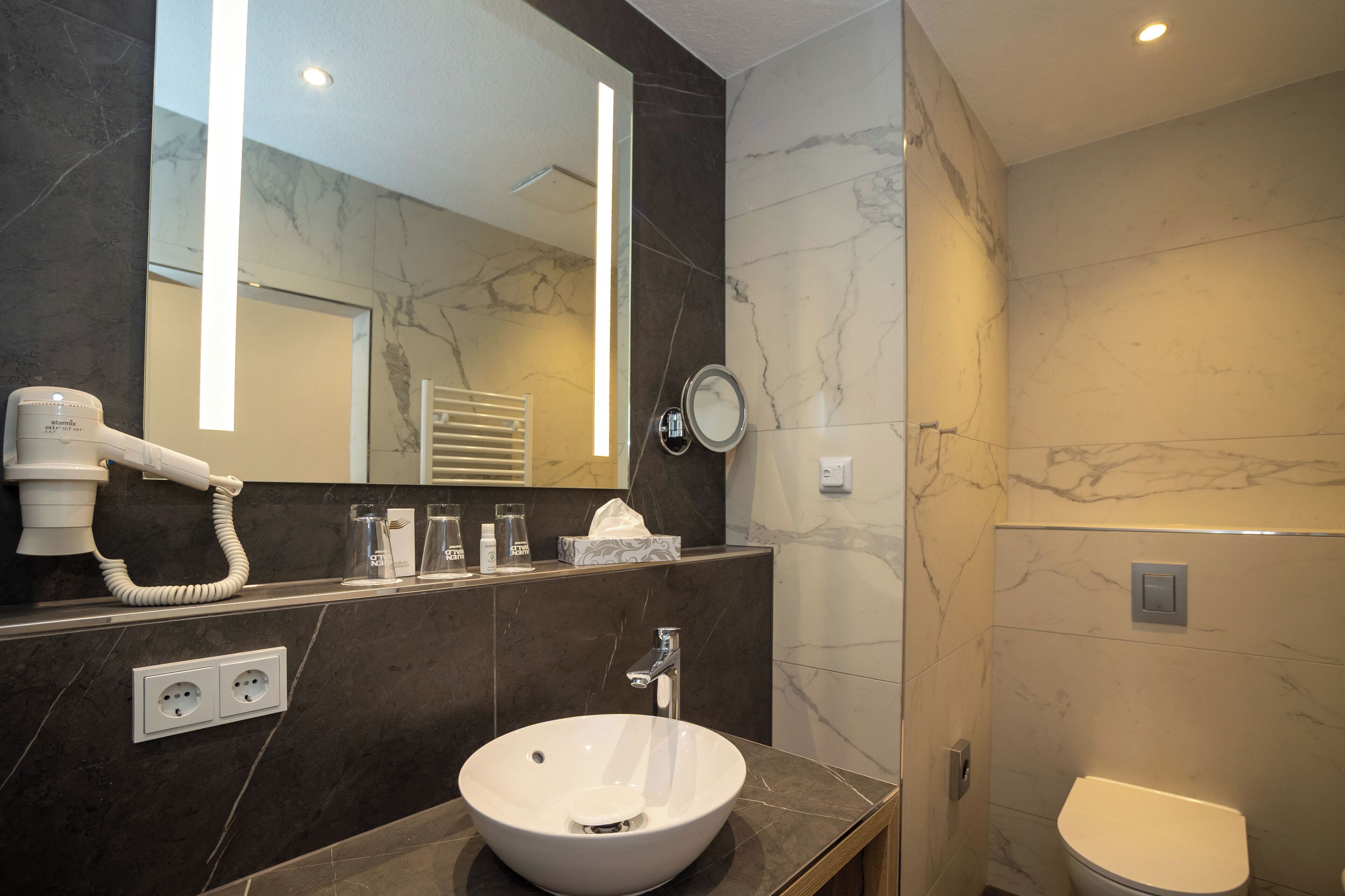 A modern bathroom with a large mirror, a sink, a toilet, and a hairdryer on the counter.