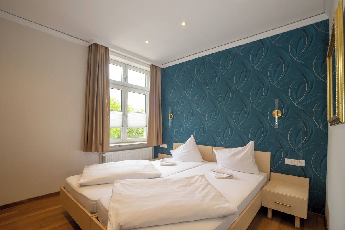Una cama doble con ropa blanca, una mesita de noche, una ventana con cortinas y una pared con diseño azul aguamarina.