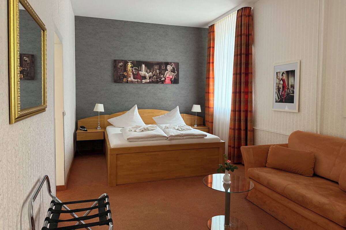 Habitación de hotel con cama, sofá, una mesita, una silla y un cuadro en la pared.