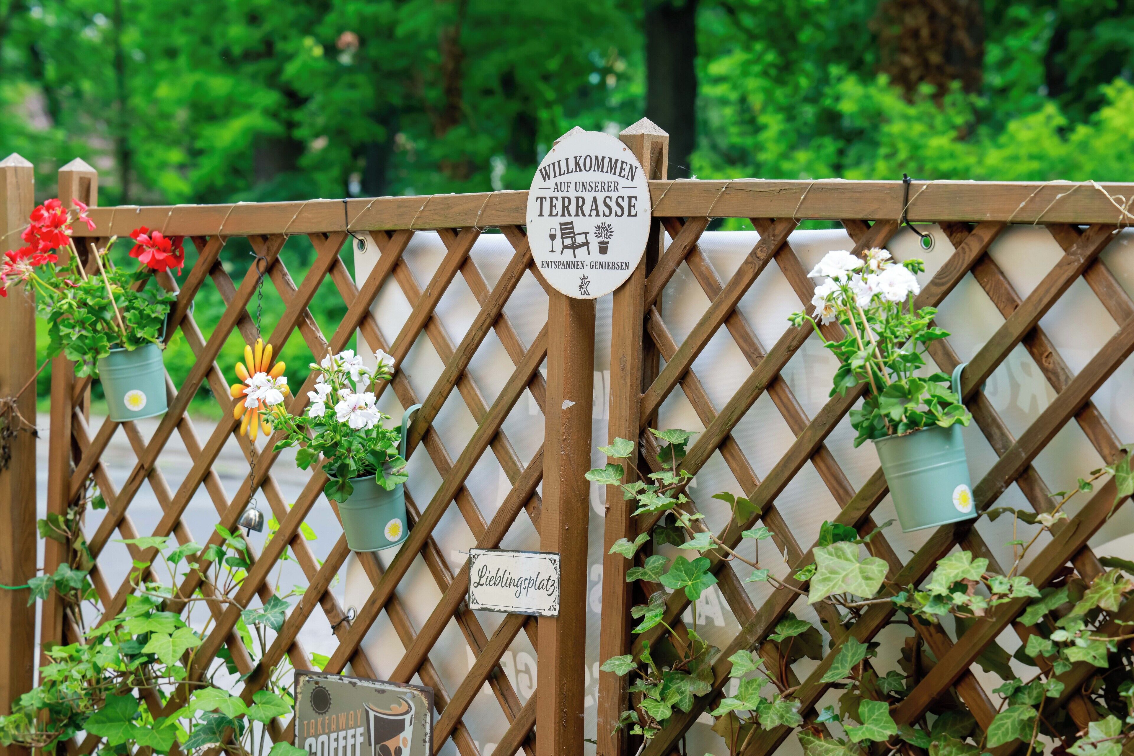 A wooden lattice fence with potted plants and signs, including one that says 'WILLKOMMEN AUF UNSERER TERRASSE' and another that says 'Lieblingsplatz'.