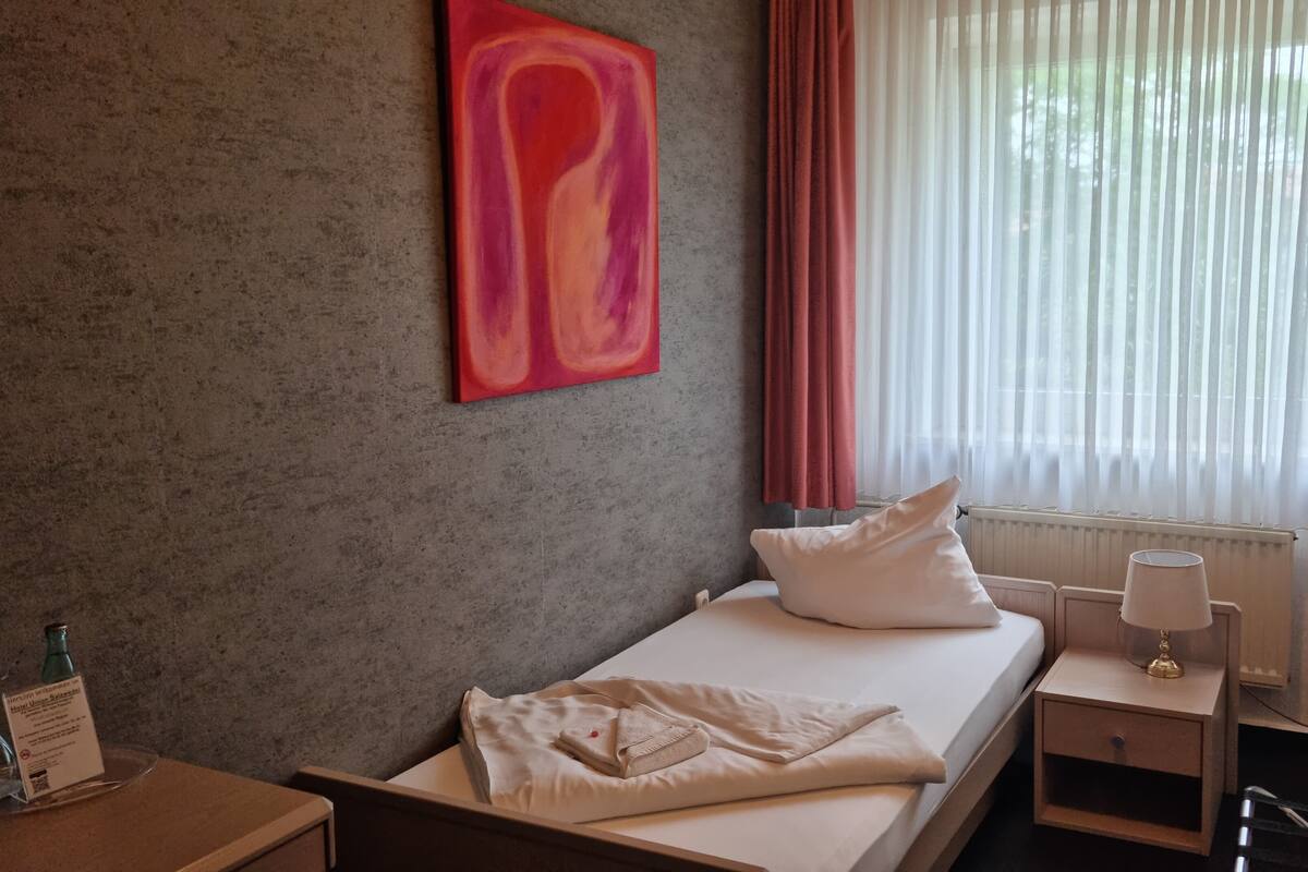 Habitación de hotel con una cama, una mesita de noche, una lámpara, una ventana con cortinas y un cuadro abstracto rojo en la pared.