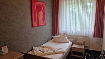 A hotel room with a bed, a nightstand, a lamp, a window with curtains, and a red abstract painting on the wall.