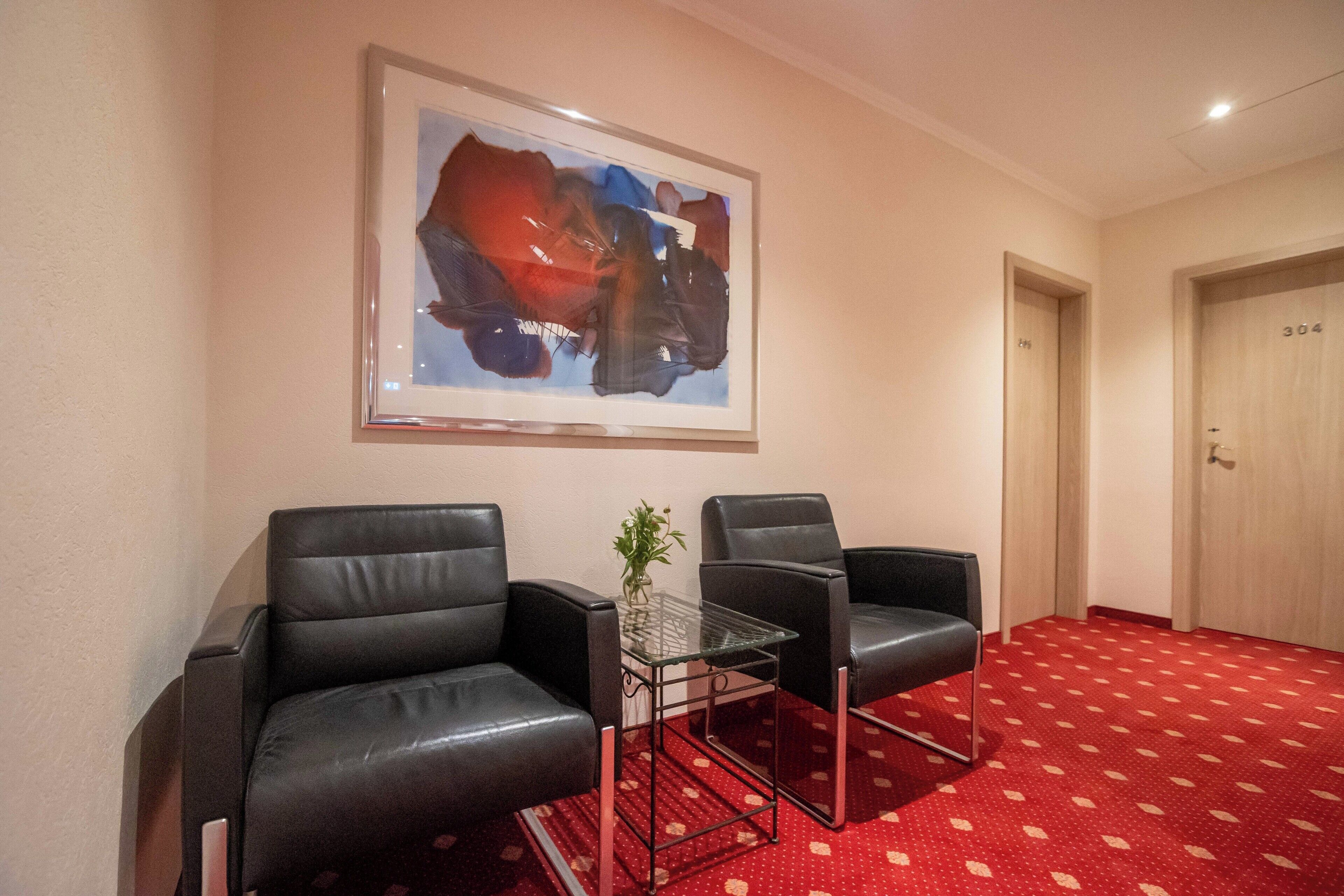A hotel room with two black leather chairs, a small glass table with a plant, a framed abstract painting on the wall, and a red patterned carpet.