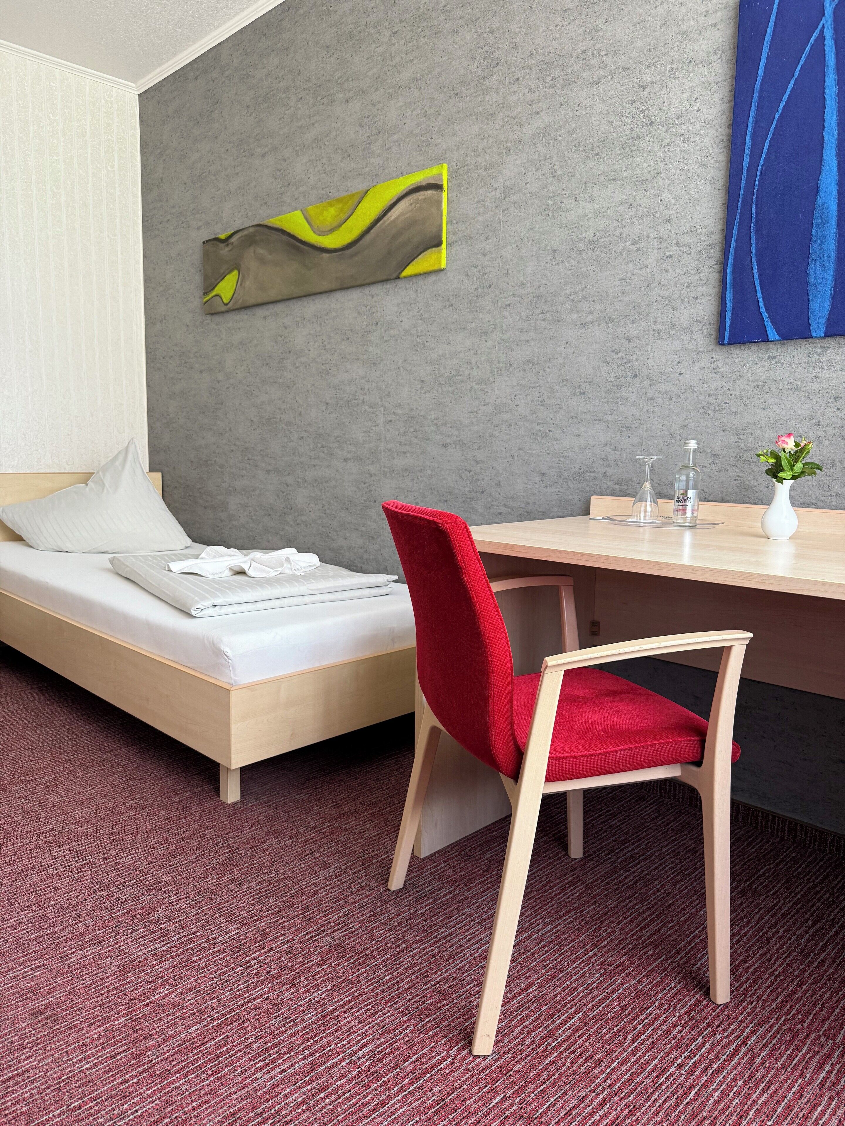 A hotel room with a bed, a desk, a red chair, a vase with a flower, and a painting on the wall.