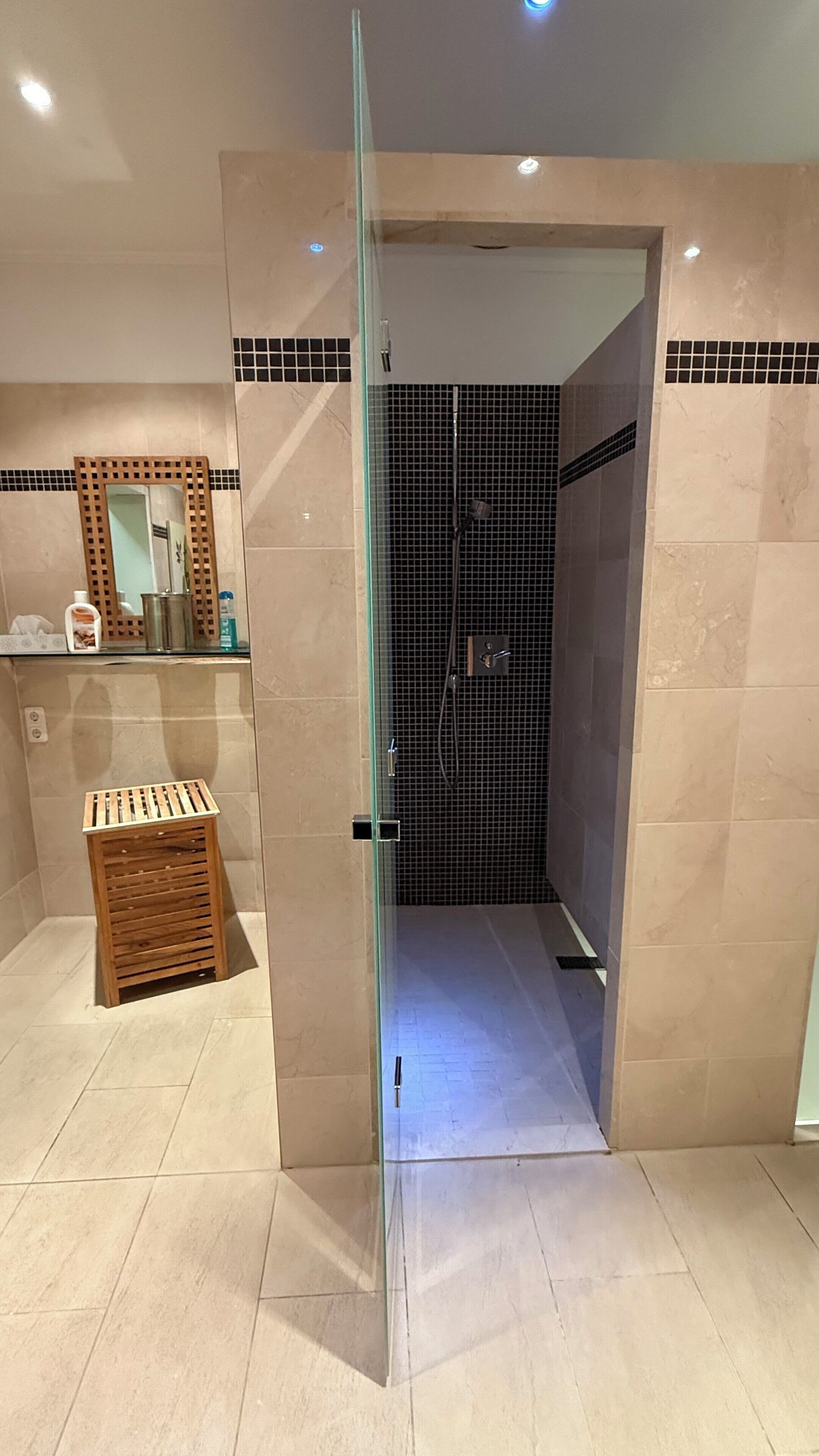 Un bagno moderno con cabina doccia in vetro, uno sgabello di legno e pavimento piastrellato.