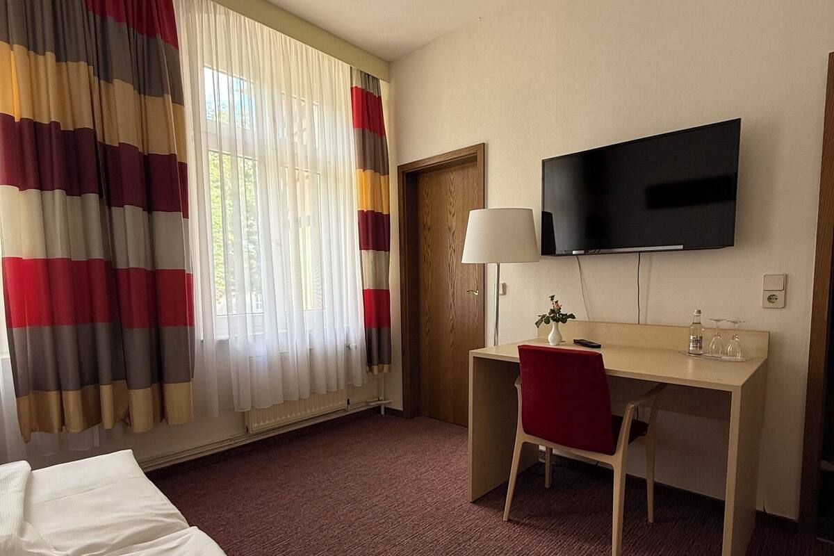 Habitación de hotel con cama, escritorio, silla, televisor y ventana con cortinas.