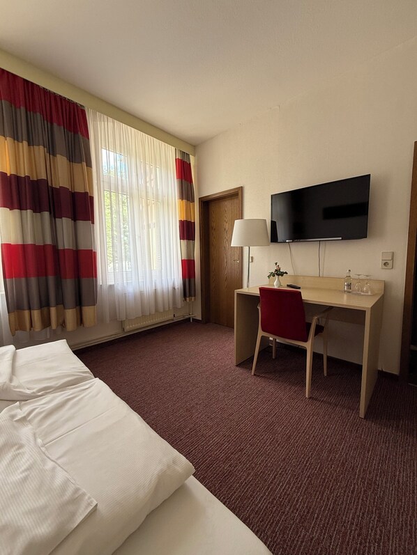Ett hotellrum med en säng, ett skrivbord, en stol, en tv och ett fönster med gardiner.