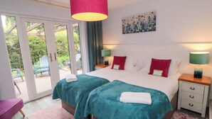 Una cama doble con ropa de cama verde aguamarina y blanca, dos almohadas rojas, una silla rosa, una mesita de noche de madera con lámpara, y un ventanal corredero que da a un espacio exterior con sillas y follaje.