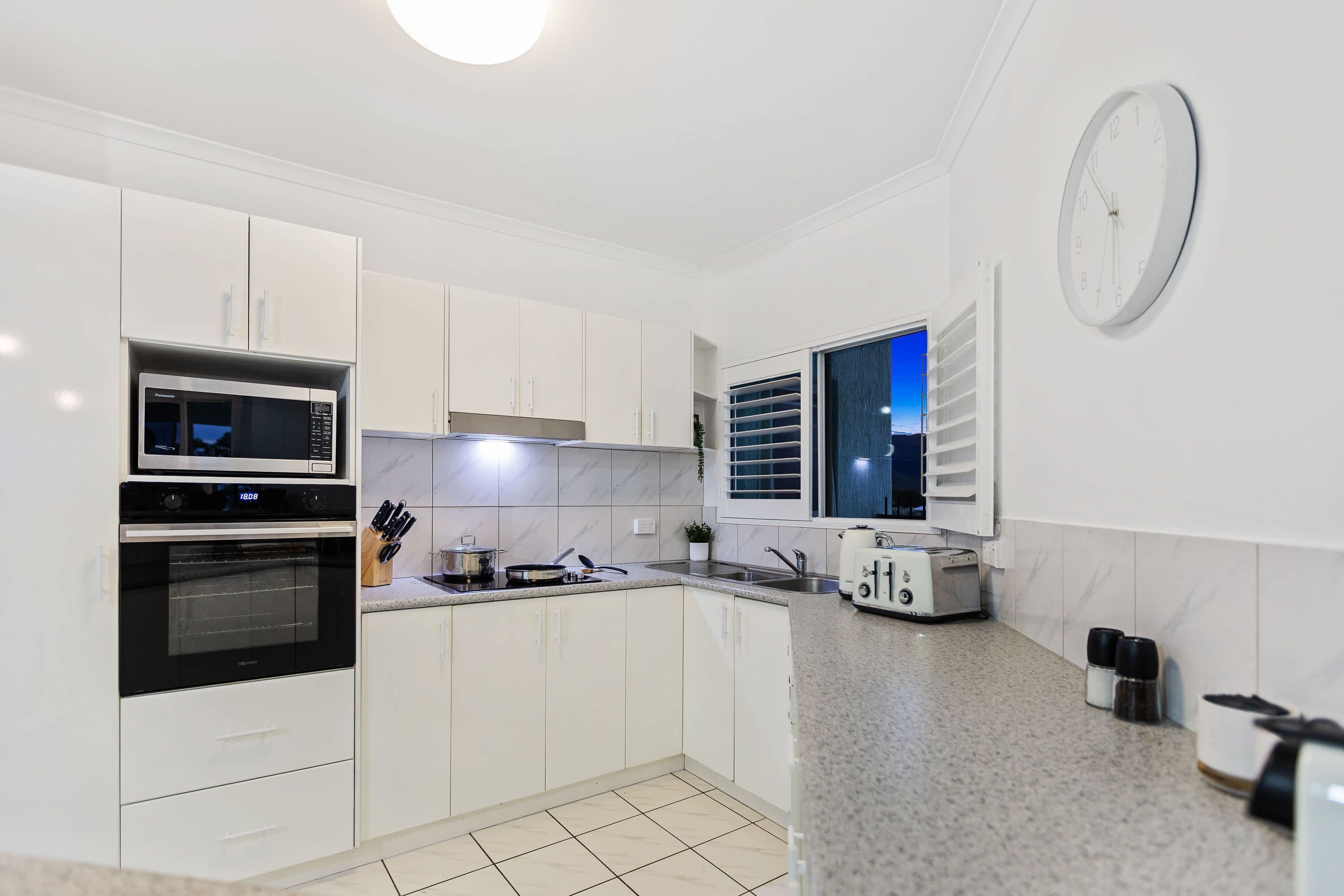 A modern kitchen with white cabinets, a microwave, an oven, a toaster, and a window with blinds.