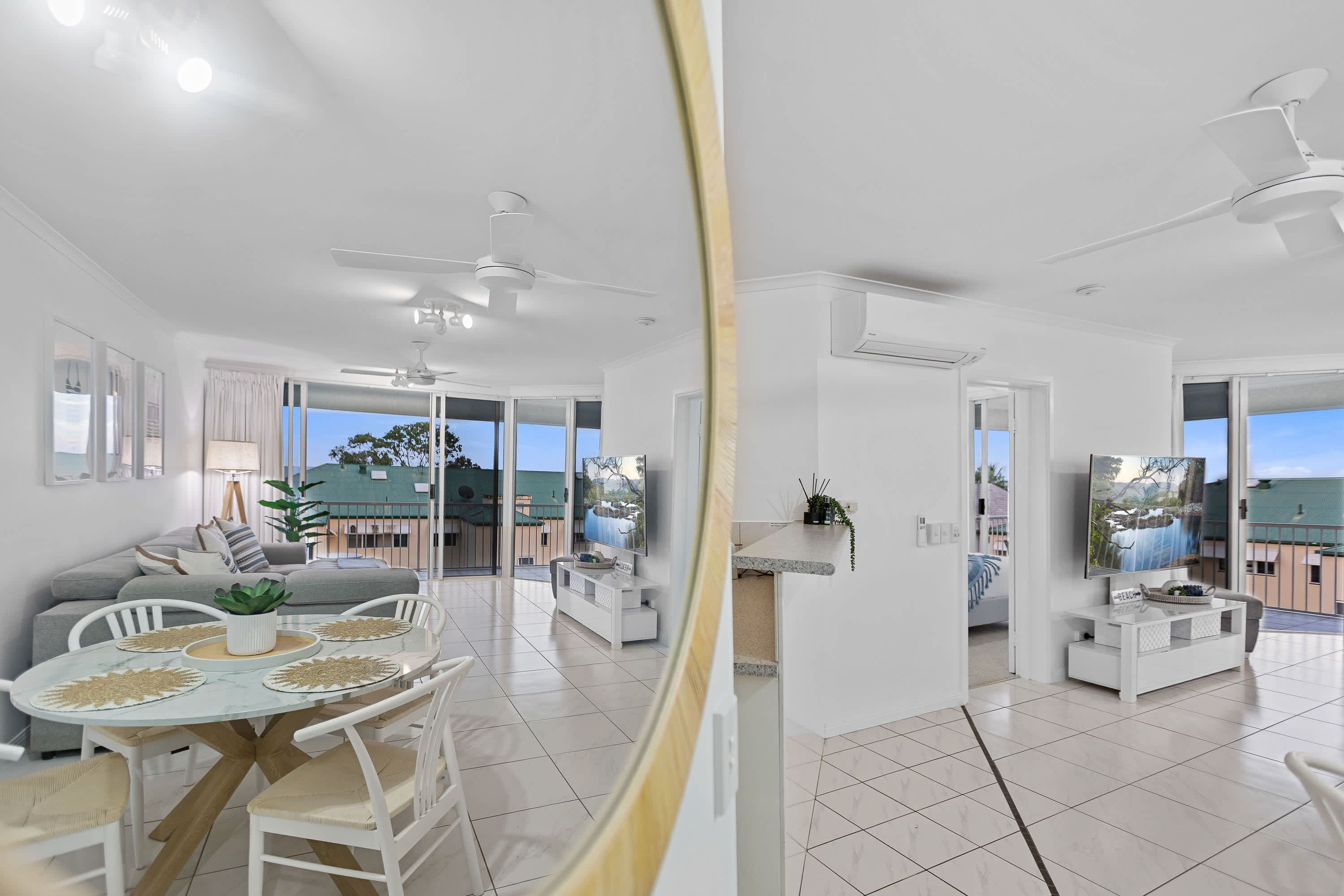 Una sala de estar moderna con comedor, muebles blancos, ventiladores de techo y ventanas amplias que ofrecen vistas al exterior.