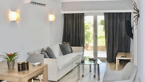 Un saló modern amb un sofà blanc, una taula de centre de vidre i una taula de menjador de fusta. Hi ha dues làmpades penjades a la paret, una planta en maceta i una vista a través de grans portes corredisses cap a una zona exterior.