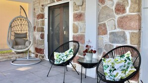 Paret exterior de pedra, una porta de vidre, una cadira penjant i dues cadires amb coixins florals.