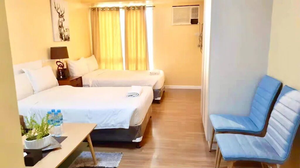 Ein Hotelzimmer mit zwei Betten, einem Schreibtisch mit Pflanze und Wasserflaschen, einem blauen Stuhl und einer Wand-Klimaanlage.