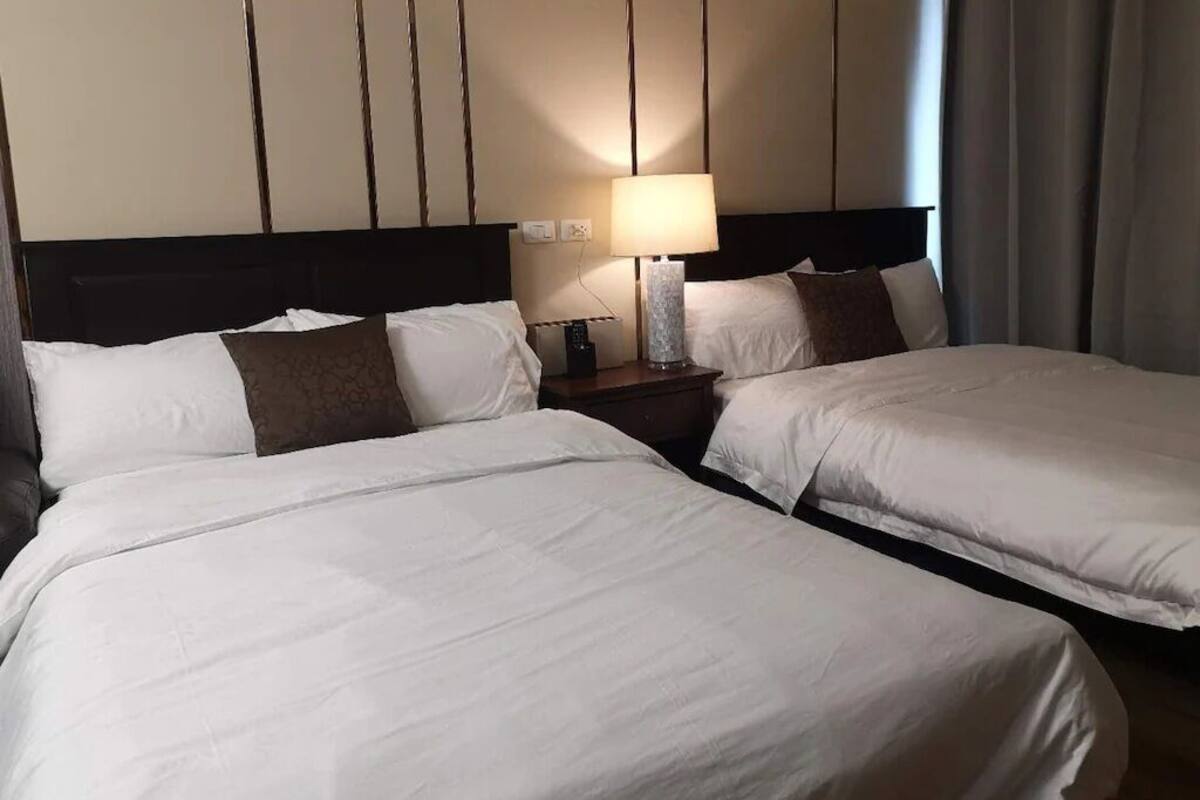 A hotel room with two beds, a nightstand with a lamp, and a view of the outside through a window.