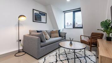 Ein modernes Wohnzimmer mit grauem Sofa, einem runden Couchtisch, einer Stehlampe und einem großen Fenster mit Blick auf Gebäude.