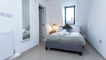 Ein Schlafzimmer mit Bett, Nachttisch, Lampe, Fenster und Spiegel.