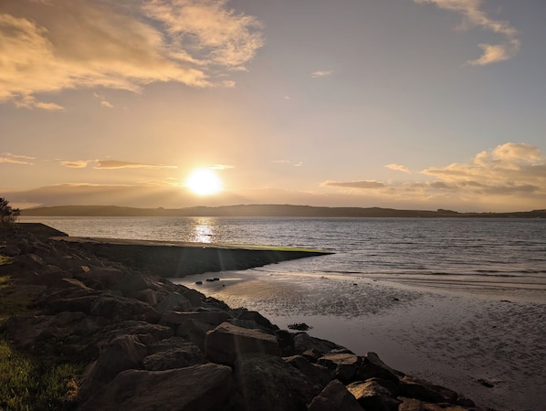 A coastal landscape with a rocky shoreline, calm water, and a distant sun setting or rising.