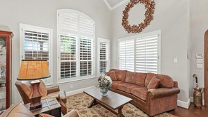 A living room with a brown sofa, a coffee table, and a lamp. There are three windows with white shutters, and a decorative wreath on the wall.