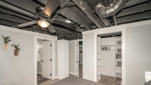 A spacious room with a high ceiling, exposed ductwork, and a ceiling fan. There are white walls, a patterned carpet, and a red chair. - Modern Cincy Home – For 6 (Cincinnati)