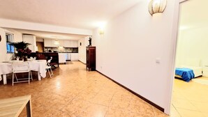 Un vaste espace de vie comprenant un coin repas avec une table dressée pour quatre personnes, une cuisine à l’arrière-plan et une chambre visible à travers une porte ouverte.