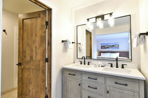 A bathroom with a wooden door, a large mirror, and a white sink with two faucets. - Pine Ridge Off-Piste Condo - Ski In, Walk to Downtown! (Breckenridge)