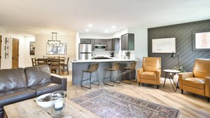 Een moderne woonkamer met een leren bank, een salontafel en een vloerkleed. Er is een keuken met een koelkast, magnetron en barstoelen.