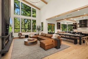 A spacious living room with a large sectional sofa, a fireplace, and large windows offering a view of the forest. - Exquisite Spa Inspired Retreat! Wet Sauna Room*Garage Game Room*Hot Tub! (Cle Elum)