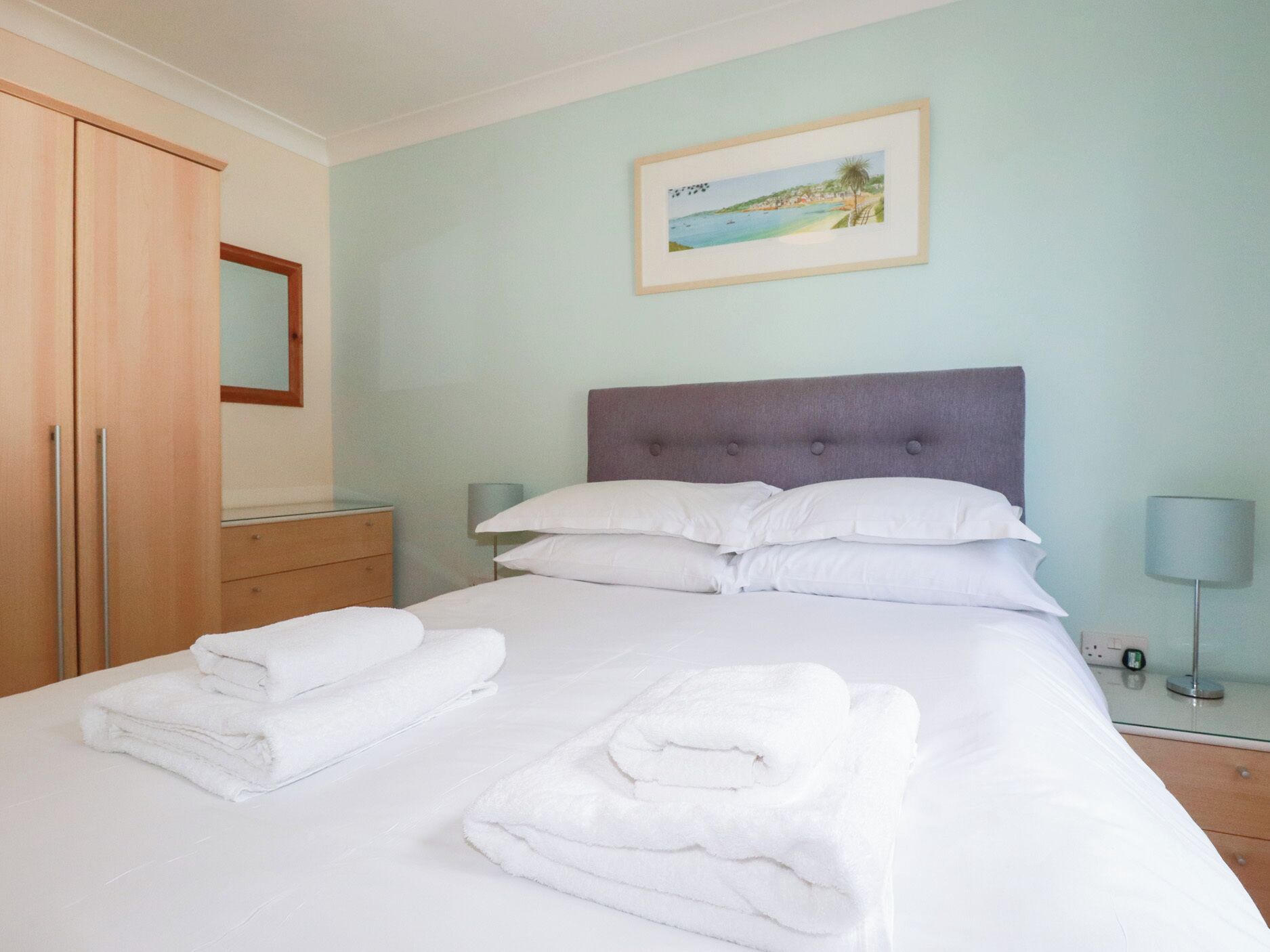 A neatly made bed with white linens and pillows, a bedside table with a lamp, a framed picture on the wall, and a wardrobe.