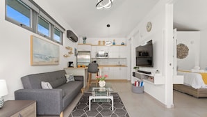 Ein modernes Wohnzimmer mit grauem Sofa, einem Glastisch und einer Küchenzeile mit weißen Schränken sowie einem Fernseher.