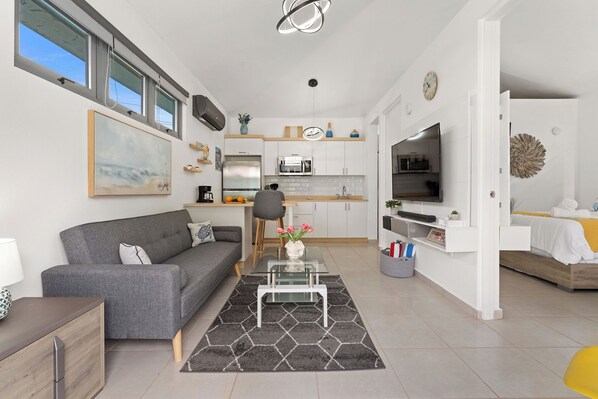 A modern living room with a grey sofa, a glass coffee table, and a kitchen area with white cabinets and a TV.