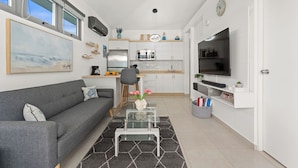 Eine moderne Küche mit weißen Schränken, einer Mikrowelle und einem Kühlschrank. Es gibt einen Essbereich mit einem grauen Sofa und einem Glastisch. Ein Fernseher ist an der Wand montiert, und ein Gemälde hängt über dem Sofa.