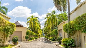 A paved street lined with palm trees and small bushes, leading to a building with a garage door. - Exotic Pool Villa Solandra in Bangtao (Choeng Thale)