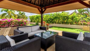 An outdoor patio with wicker furniture, a glass-top table, and a view of a lush garden with palm trees and flowering plants. - Exotic Pool Villa Solandra in Bangtao (Choeng Thale)