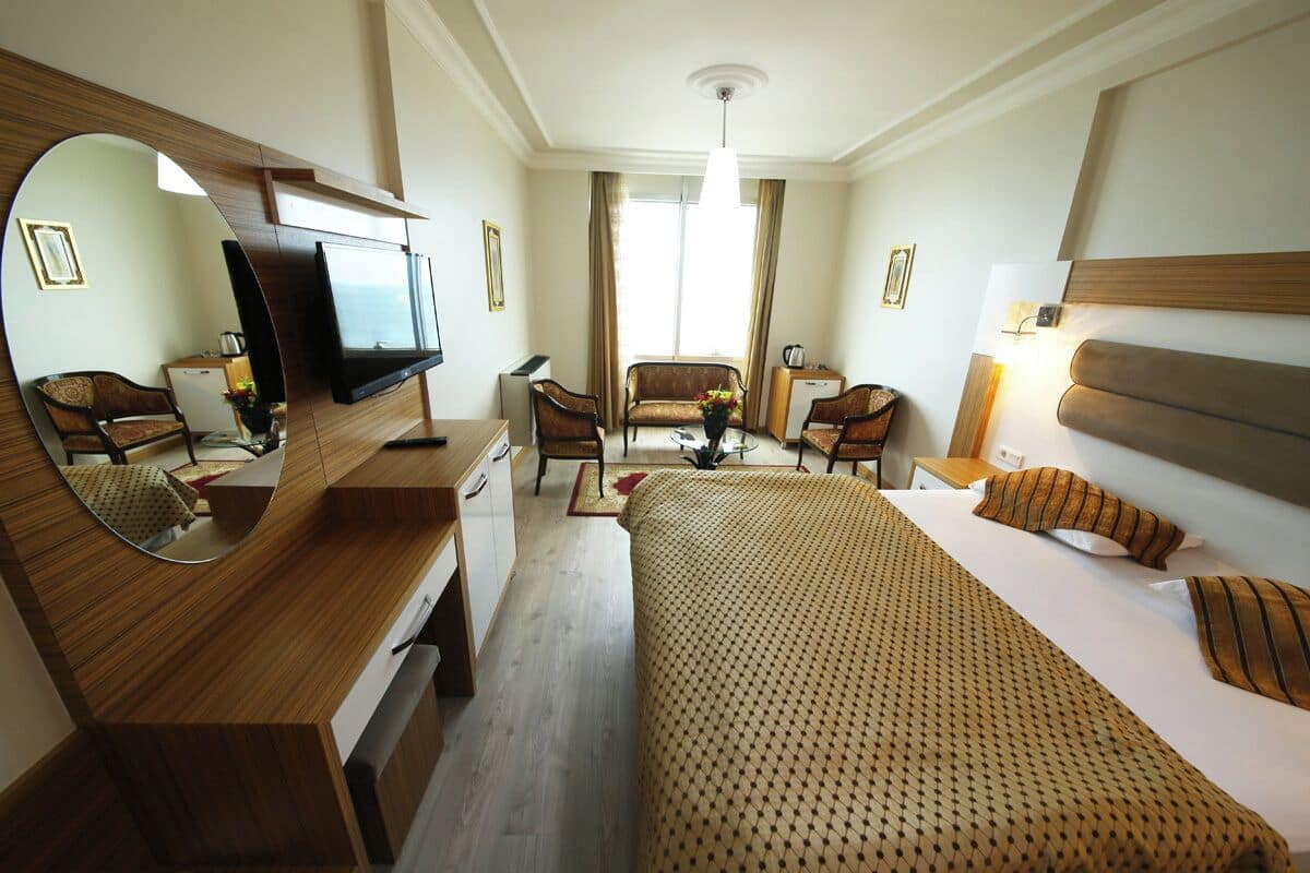 Quarto de hotel com cama, escrivaninha com televisão, poltrona, sofá e espelho.