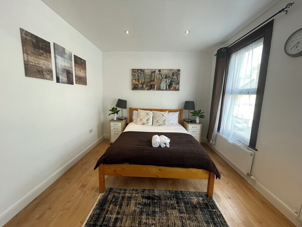A bedroom with a wooden bed, two bedside tables with lamps, a large window with curtains, and a painting on the wall.