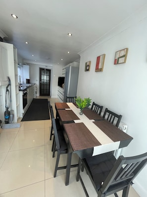 A modern kitchen with a central island, a dining area with a long table and chairs, and a hallway leading to a door. - Beautiful house with 5 bedrooms and Living Room! (London)
